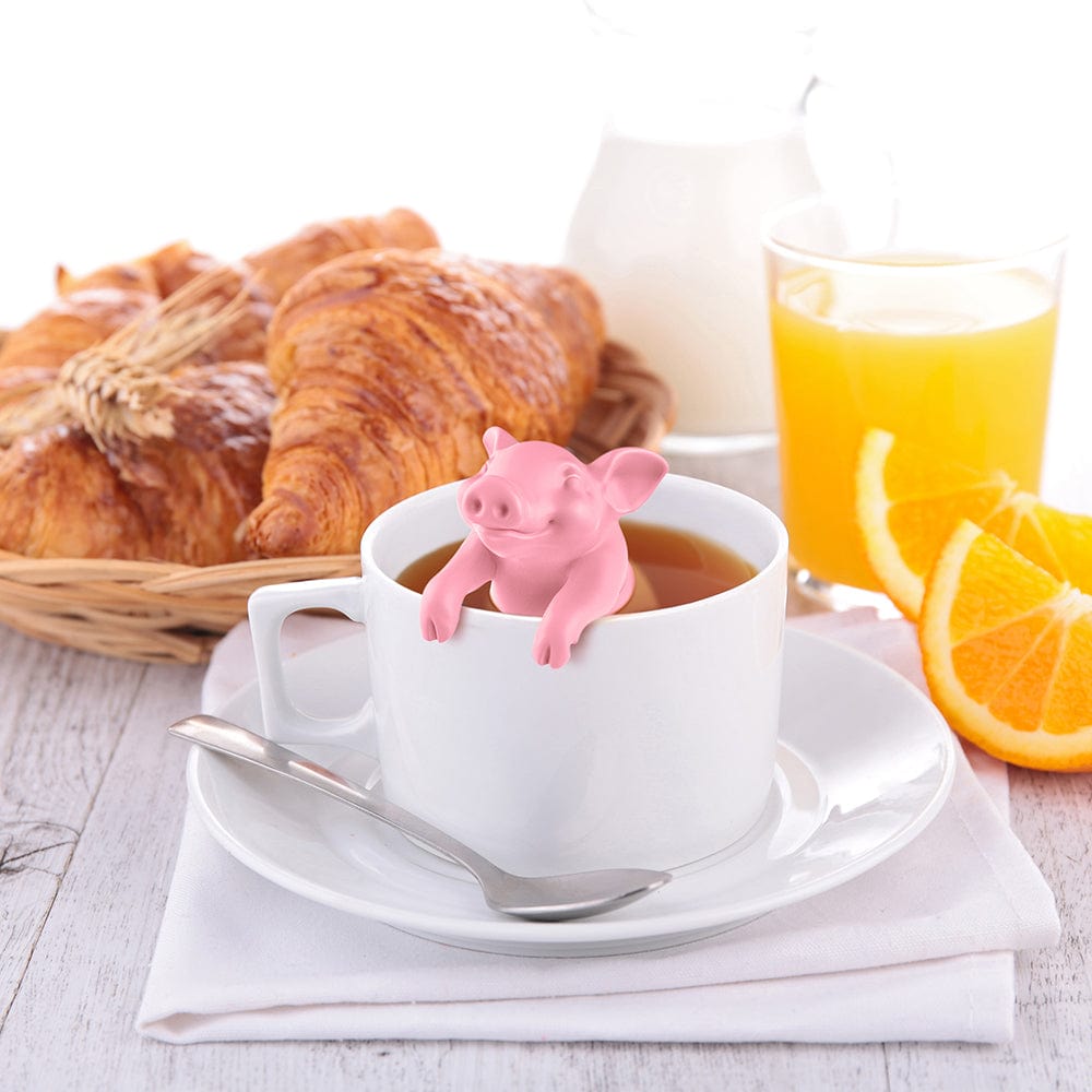 A silicone tea infuser that looks like a pink pig. Placed in a cup of tea and surrounded by croissants, oranges, orange juice, and cream