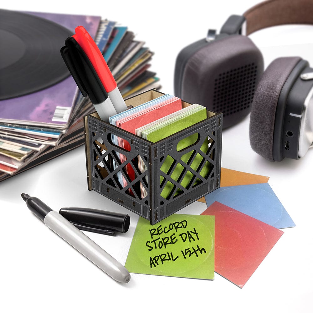 A small milk crate caddy for your desk. The crate is holding markers and notecards that look like vinyl album covers in 4 colors: orange, blue, green, and red. Behind the milk crate there is a stack of records and large headphones. There is a green notecard with handwriting on it that says " Record Store Day, April 15th" 