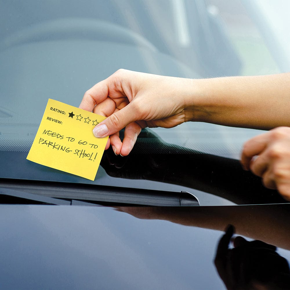 A hand placing a yellow sticky note on a car windshield. The note shows 1-star and "needs to go to parking school"