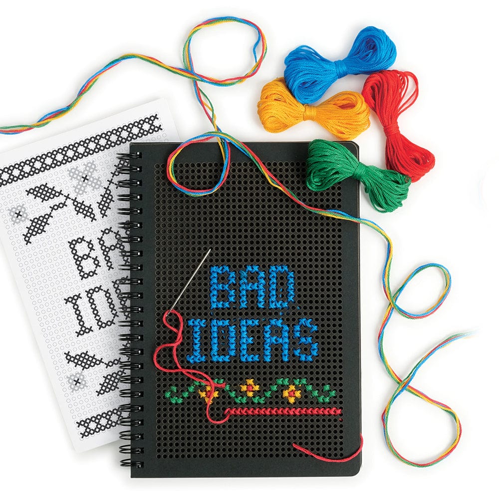 A black journal with a cross stitched cover that says "Bad Ideas". The journal is surrounded by 4 colors of embroidery thread (blue, green, red, and yellow), a black and white pattern booklet, and a metal needle.