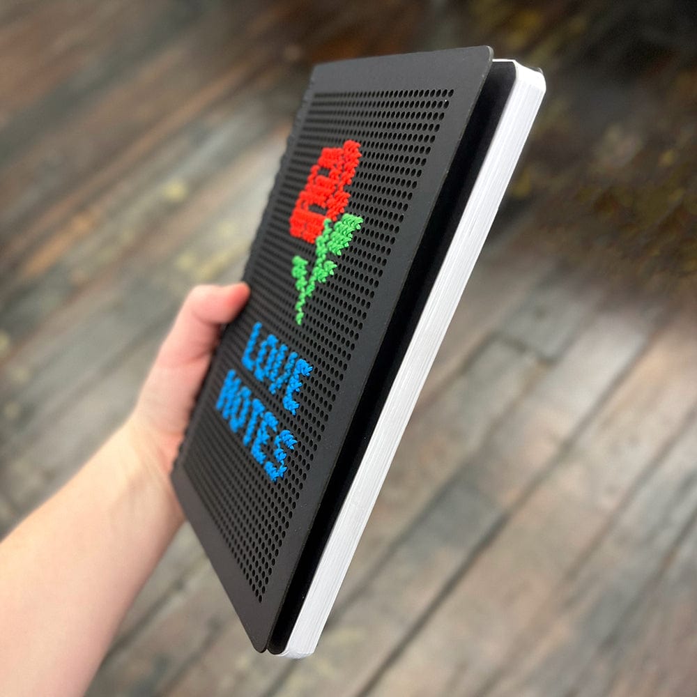 A hand holding a black journal with a cross stitched cover that says "Love Notes" and shows an image of a rose